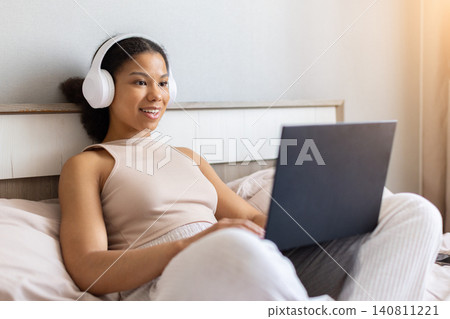 Happy black woman sitting in bed wearing headphones and using laptop 140811221