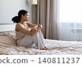 Thoughtful black woman sitting on bed hugging knees and looking toward window 140811237