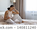 Black woman sitting on bed, holding glass of water and touching head 140811240