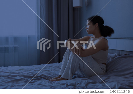 Black woman is sitting on bed at night and looking toward window Black woman is sitting on bed at night and looking toward window 140811246