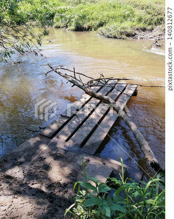 wooden bridge or flooring with dry tree branches lies in the water near the muddy bank of a small forest river in summer wooden bridge or flooring with dry tree branches lies in the water near the muddy bank of a small forest river in summer 140812677