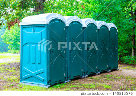 Several portable restrooms stand in line on dirt path in park surrounded by trees. Several portable restrooms stand in line on dirt path in park surrounded by trees. 140813576