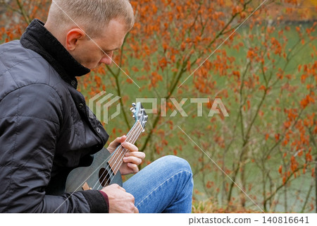 Man plays ukulele while spending time outdoors in autumnal natural setting. Musician holds small string instrument and performs music in relaxed atmosphere surrounded by greenery and open space Man plays ukulele while spending time outdoors in autumnal natural setting. Musician holds small string instrument and performs music in relaxed atmosphere surrounded by greenery and open space 140816641