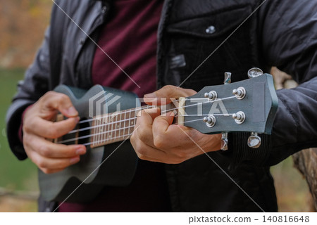 Man plays ukulele while spending time outdoors in autumnal natural setting. Musician holds small string instrument and performs music in relaxed atmosphere surrounded by greenery and open space 140816648