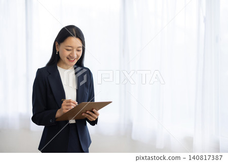 A young Japanese businesswoman smiles as she takes notes in a binder by the window. A young Japanese businesswoman smiles as she takes notes in a binder by the window. 140817387