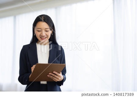 A young Japanese businesswoman smiles as she takes notes in a binder by the window. A young Japanese businesswoman smiles as she takes notes in a binder by the window. 140817389