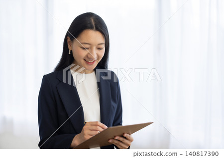 A young Japanese businesswoman smiles as she takes notes in a binder by the window. A young Japanese businesswoman smiles as she takes notes in a binder by the window. 140817390
