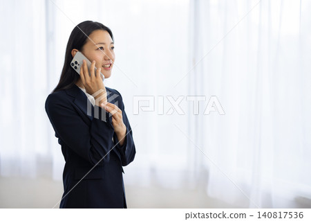 A young Japanese businesswoman smiles and talks on her smartphone by the window. A young Japanese businesswoman smiles and talks on her smartphone by the window. 140817536
