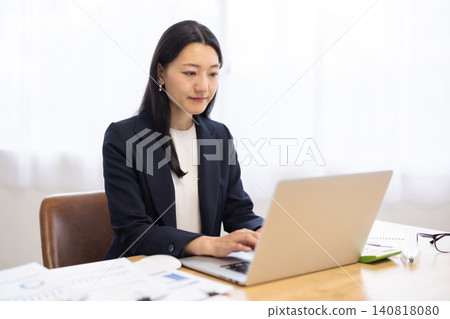 A young Japanese businesswoman working on her computer by a bright window. 140818080