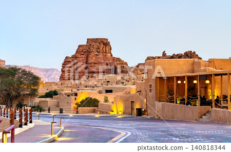 View of the historic mud brick architecture and sandstone mountains in AlUla Old Town, Saudi Arabia, featuring a modern walkway with ambient lighting at dusk. 140818344