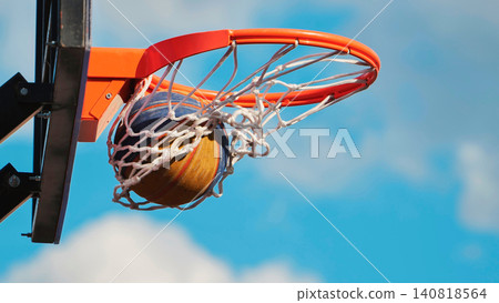 Basketball going through an orange hoop and net, scoring a point against a bright blue sky Basketball going through an orange hoop and net, scoring a point against a bright blue sky 140818564