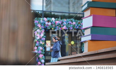 Large colorful books stacked on a wooden bench with a blurred person and floral arch in an urban setting Large colorful books stacked on a wooden bench with a blurred person and floral arch in an urban setting 140818565