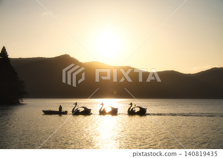 The silhouettes of swan boats gliding across the lake at sunset, shimmering in golden light, and the beautiful sunlit road. The silhouettes of swan boats gliding across the lake at sunset, shimmering in golden light, and the beautiful sunlit road. 140819435