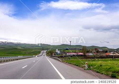 Serene rural highway with open sky, green fields, and distant village houses 140819520
