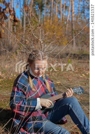 Beautiful blonde girl playing guitar ukulele and singing relaxing enjoying outdoors on sunny autumn day. Hippie woman in boho style training playing musical instrument. Fall leaves Beautiful blonde girl playing guitar ukulele and singing relaxing enjoying outdoors on sunny autumn day. Hippie woman in boho style training playing musical instrument. Fall leaves 140821537
