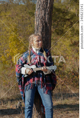 Beautiful blonde girl playing guitar ukulele and singing relaxing enjoying outdoors on sunny autumn day. Hippie woman in boho style training playing musical instrument. Fall leaves Beautiful blonde girl playing guitar ukulele and singing relaxing enjoying outdoors on sunny autumn day. Hippie woman in boho style training playing musical instrument. Fall leaves 140821541
