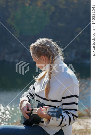 Blonde woman playing guitar ukulele outdoors in autumn forest. Concept of sound therapy, mental health and wellness rituals. Calmness tranquility audio-sensory practices. Aura farming Blonde woman playing guitar ukulele outdoors in autumn forest. Concept of sound therapy, mental health and wellness rituals. Calmness tranquility audio-sensory practices. Aura farming 140821542