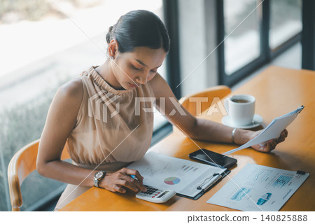 Top view of Asian businesswoman calculating financial data with reports, graphs, smartphone, and coffee on cafe table, ideal for budgeting, analytics, accounting, and planning workflow. 140825888