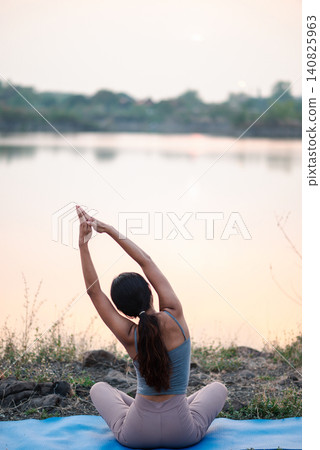 Back view Asian woman practicing seated yoga side stretch on blue mat by a calm lake at sunset, peaceful outdoor wellness, mindful breathing, flexibility training, nature relaxation and self care. Back view Asian woman practicing seated yoga side stretch on blue mat by a calm lake at sunset, peaceful outdoor wellness, mindful breathing, flexibility training, nature relaxation and self care. 140825963