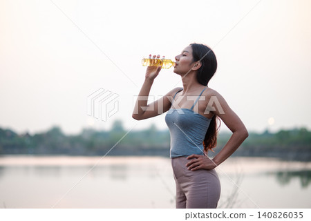 Asian woman drinking water after outdoor yoga beside a calm lake at sunset, healthy wellness lifestyle, hydration, mindful recovery, fresh air, peaceful nature fitness and active self care. Asian woman drinking water after outdoor yoga beside a calm lake at sunset, healthy wellness lifestyle, hydration, mindful recovery, fresh air, peaceful nature fitness and active self care. 140826035