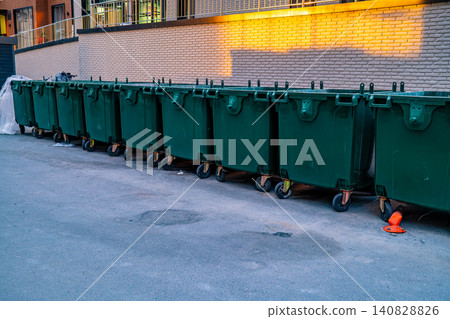 Row of green large wheeled garbage containers lined up on city street at sunset, waste management and urban sanitation concept Row of green large wheeled garbage containers lined up on city street at sunset, waste management and urban sanitation concept 140828826