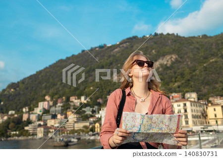 Happy blonde woman traveler searching for direction with city map against mountain lake Happy blonde woman traveler searching for direction with city map against mountain lake 140830731