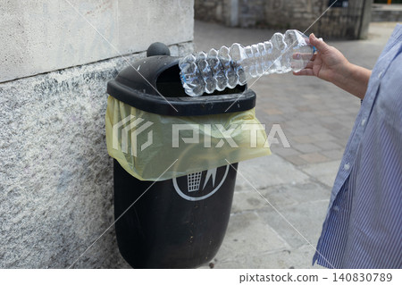 Person throwing empty plastic water bottle into public garbage bin outdoors 140830789