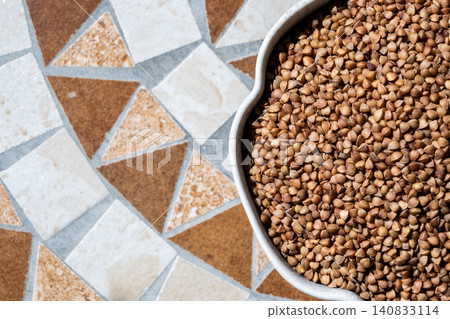 Gray bowl with uncooked roasted buckwheat on the stone table for healthy food. Cooking concept. Top view. Gray bowl with uncooked roasted buckwheat on the stone table for healthy food. Cooking concept. Top view. 140833114
