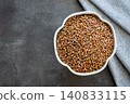 Gray bowl with uncooked roasted buckwheat on the black stone table with napkin for healthy food. Cooking concept. Top view. Space for text. 140833115