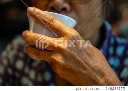 An elderly person with silver hair holds a white porcelain teacup in both hands and sips tea at a teahouse gathering. An elderly person with silver hair holds a white porcelain teacup in both hands and sips tea at a teahouse gathering. 140833155