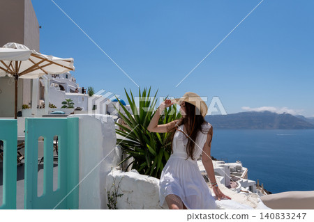 Beautiful young woman in white dress and straw hat sitting on terrace wall in Santorini Greece Beautiful young woman in white dress and straw hat sitting on terrace wall in Santorini Greece 140833247