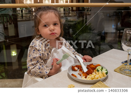 Little girl looking down at food in restaurant high chair, cute toddler holding plastic fork and knife trying to pick up chicken nugget, children menu and parenting concept Little girl looking down at food in restaurant high chair, cute toddler holding plastic fork and knife trying to pick up chicken nugget, children menu and parenting concept 140833306