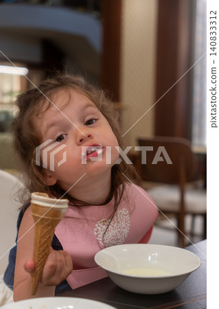 Vertical shot of cute little girl with dirty face eating ice cream in restaurant, funny toddler holding waffle cone at table, messy kid with milk mustache after dessert Vertical shot of cute little girl with dirty face eating ice cream in restaurant, funny toddler holding waffle cone at table, messy kid with milk mustache after dessert 140833312