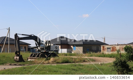 Construction of a private home outside the city. A yellow excavator stands on the site near an unfinished building with a dark roof. A trailer, some blocks, and a dirt road are visible nearby. Construction of a private home outside the city. A yellow excavator stands on the site near an unfinished building with a dark roof. A trailer, some blocks, and a dirt road are visible nearby. 140833654