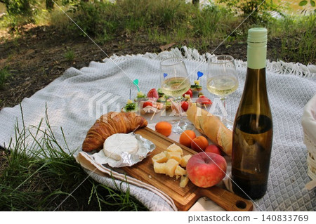 Picnic setup featuring assorted cheeses, croissants, fruits, and white wine on a blanket by a serene lakeside surrounded by greenery and natural light Picnic setup featuring assorted cheeses, croissants, fruits, and white wine on a blanket by a serene lakeside surrounded by greenery and natural light 140833769