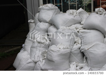 Pile of white fabric sacks stacked against a building, showcasing the texture and shape of the bags in an outdoor setting with a blurred background Pile of white fabric sacks stacked against a building, showcasing the texture and shape of the bags in an outdoor setting with a blurred background 140833780