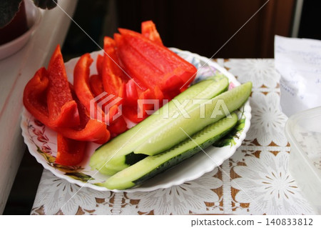 Freshly sliced red bell peppers and cucumbers arranged on a decorative plate with floral design, placed on a patterned tablecloth indoors Freshly sliced red bell peppers and cucumbers arranged on a decorative plate with floral design, placed on a patterned tablecloth indoors 140833812