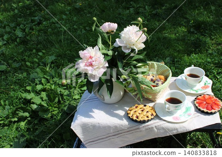 Fresh peonies in a white vase beside a basket of pastries and two cups of coffee on a table in a green garden setting with sunlight illuminating the scene Fresh peonies in a white vase beside a basket of pastries and two cups of coffee on a table in a green garden setting with sunlight illuminating the scene 140833818