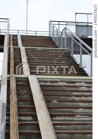Metal staircase with weathered steps leading upward, surrounded by railings and concrete structure under a cloudy sky in an urban environment Metal staircase with weathered steps leading upward, surrounded by railings and concrete structure under a cloudy sky in an urban environment 140833825