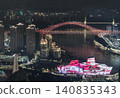 Chongqing awakens in a breathtaking display of architectural elegance and glowing currents. A majestic, crimson-lit arch bridge spans gracefully across the dark water, reflecting softly upon the river 140835343