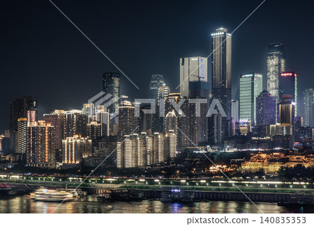 Chongqing nocturnal cityscape unfolds as a towering amphitheater of light. Modern skyscrapers pierce the dark sky with crisp white, amber, and cool aquamarine tones. Layered high-rises stack densely 140835353