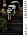Two figures under pale, luminous umbrellas ascend the dark, rain-slicked stone steps of Luohan Temple in Chongqing, their quiet ascent framed by ancient temple architecture and vibrant green foliage. 140835386