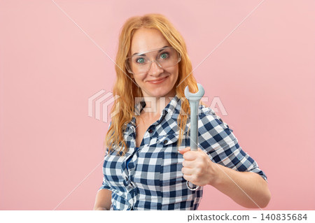 Smiling woman wearing safety goggles and a plaid shirt, confidently holding a wrench and looking at the camera against a pink background, representing skill and readiness for work Smiling woman wearing safety goggles and a plaid shirt, confidently holding a wrench and looking at the camera against a pink background, representing skill and readiness for work 140835684