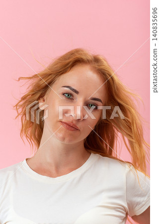 Confident young woman with flowing red hair and green eyes expressing a charming attitude while standing against a solid pink background Confident young woman with flowing red hair and green eyes expressing a charming attitude while standing against a solid pink background 140835696