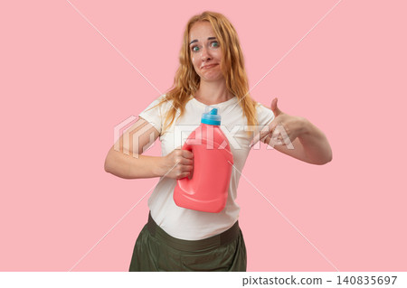 Young woman holding a large container of laundry detergent, looking confused at the camera and pointing downwards, featuring household chores concept Young woman holding a large container of laundry detergent, looking confused at the camera and pointing downwards, featuring household chores concept 140835697