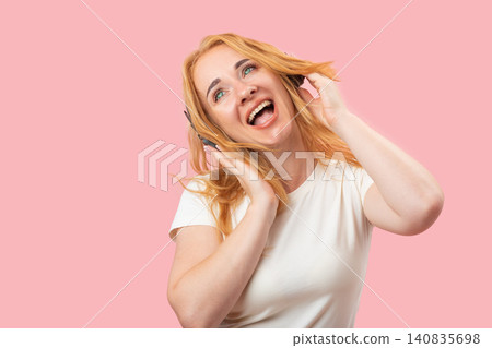 Smiling young woman with blonde hair listening to music through black headphones, expressing joy and energy while looking upwards against a pink background Smiling young woman with blonde hair listening to music through black headphones, expressing joy and energy while looking upwards against a pink background 140835698