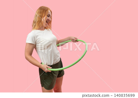 Happy young woman engaging in fitness exercise, holding a hula hoop and smiling with joy, promoting healthy lifestyle, energy, and fun on a pink background 140835699