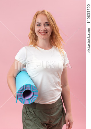 Smiling red haired woman holding a rolled blue yoga mat, ready for a fitness activity and promoting wellness, exercise, and an active lifestyle against a vibrant pink studio background Smiling red haired woman holding a rolled blue yoga mat, ready for a fitness activity and promoting wellness, exercise, and an active lifestyle against a vibrant pink studio background 140835700