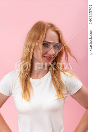 Young redhead woman wearing clear protective eyewear and a white t shirt, standing confidently with a smile against a solid pink background 140835701