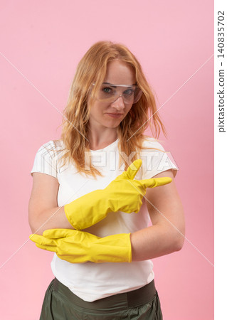 Woman wearing yellow rubber gloves and protective glasses looking at the camera while confidently pointing her gloved hand to the right against a pink background Woman wearing yellow rubber gloves and protective glasses looking at the camera while confidently pointing her gloved hand to the right against a pink background 140835702